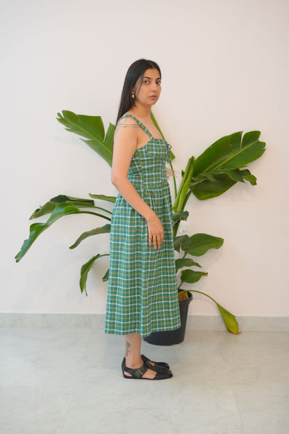 Woman in a green checkered dress standing next to a large plant against a white wall.