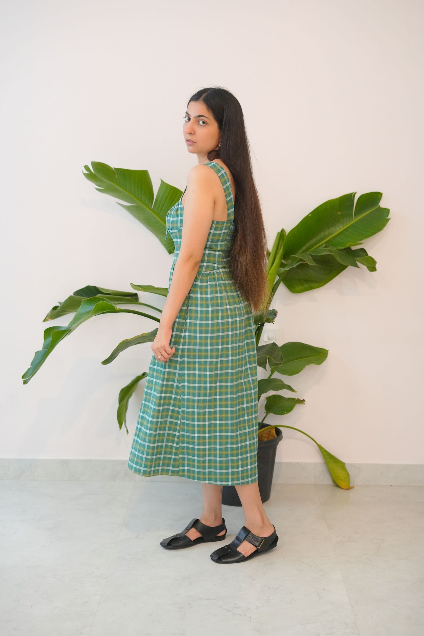 Woman in a green checkered dress standing next to a large plant against a white wall.