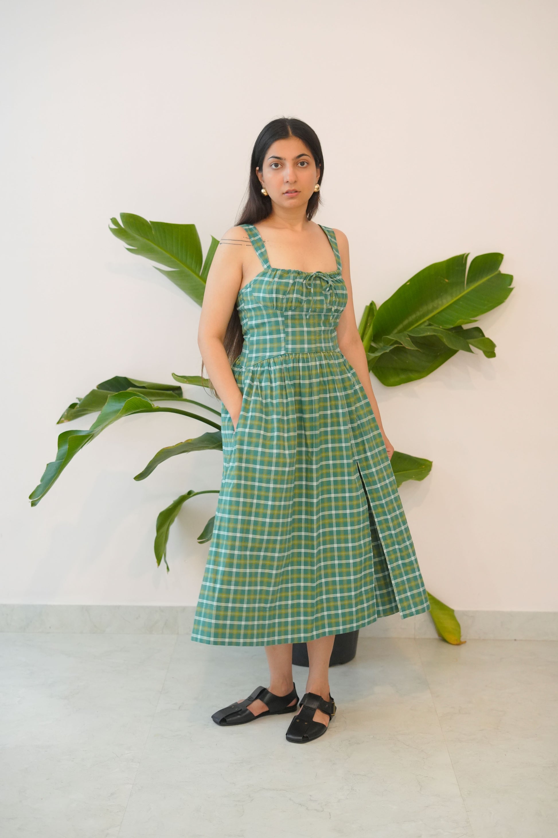 Woman wearing a green checkered dress standing between two large green leaves against a white wall.