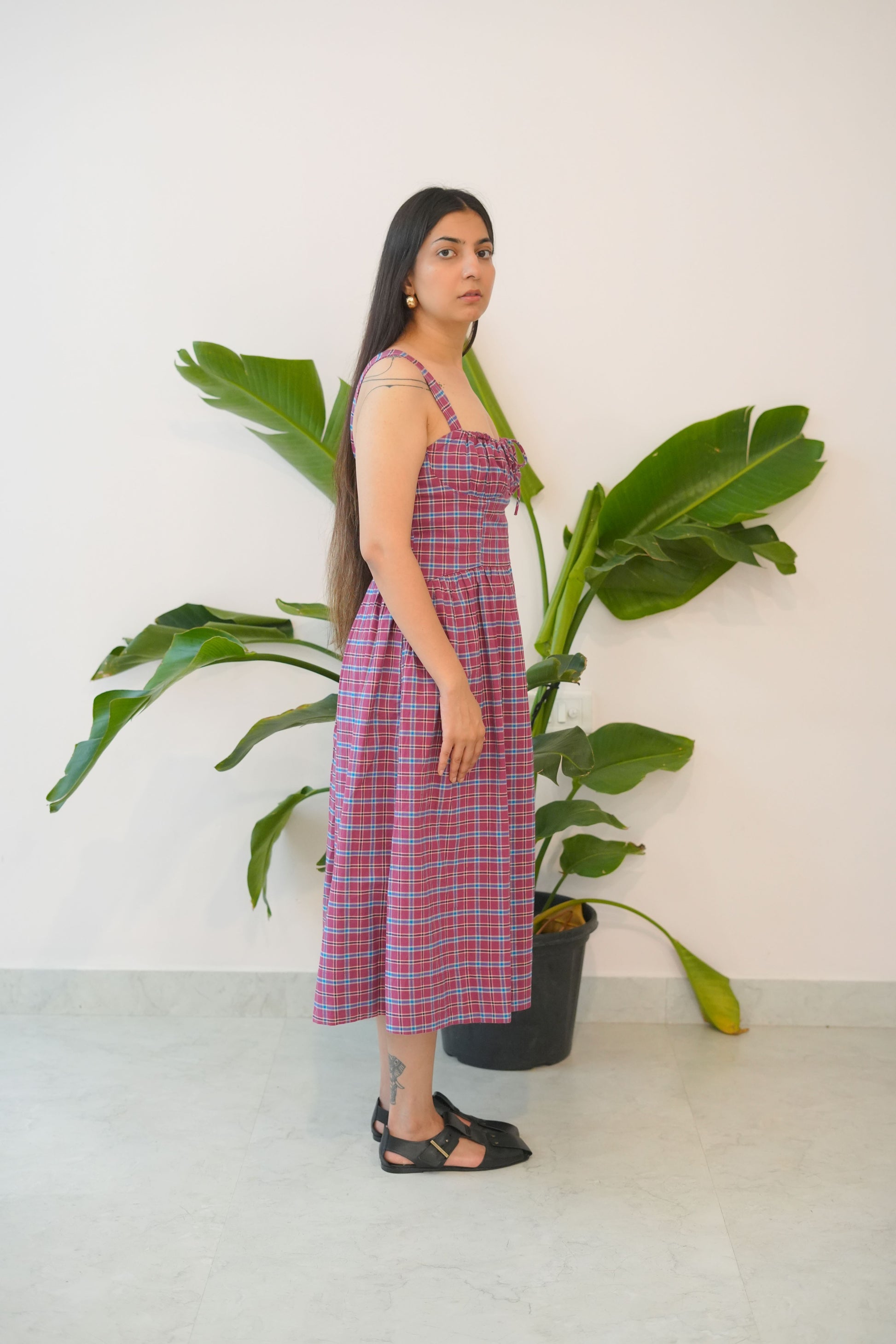 Woman wearing a red and white checkered dress standing next to a large green plant indoors.