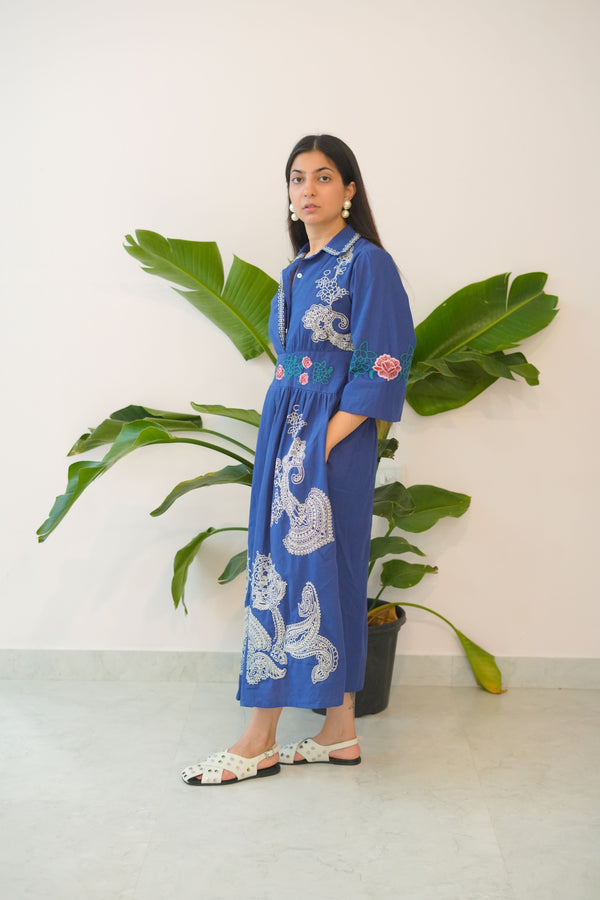 Woman in a blue dress with white embroidery lying on a white floor next to a green plant.