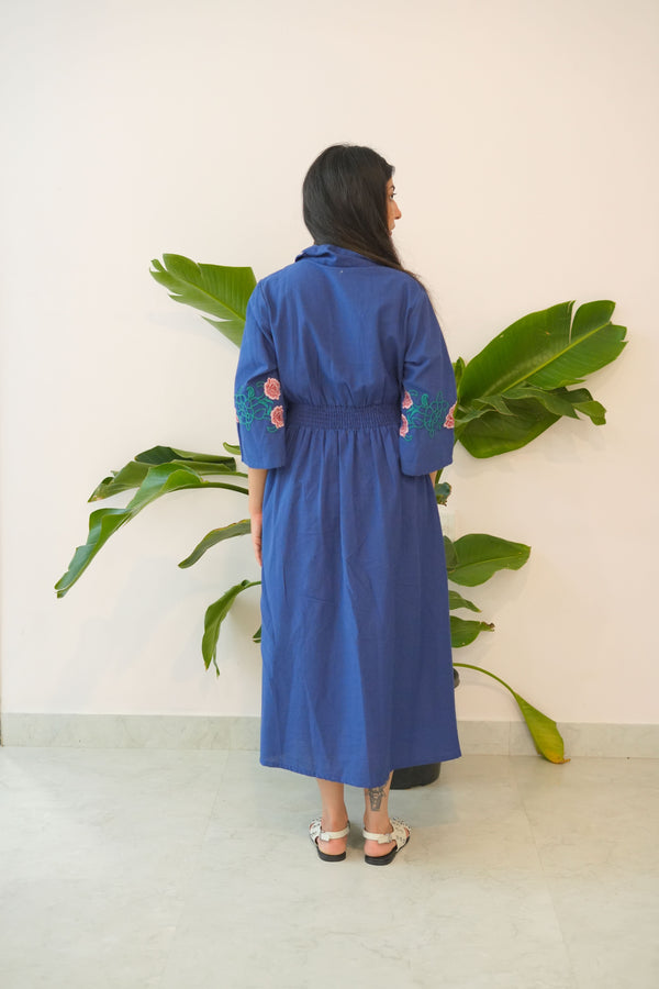 Person wearing a blue dress standing in front of a plant with a white wall background
