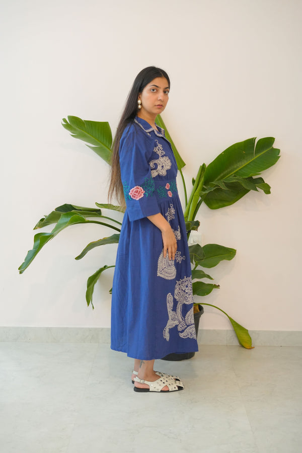 Woman in a blue embroidered dress standing next to a large green plant indoors.