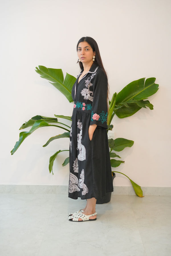 Person wearing a black dress with floral patterns standing next to a large green plant indoors.