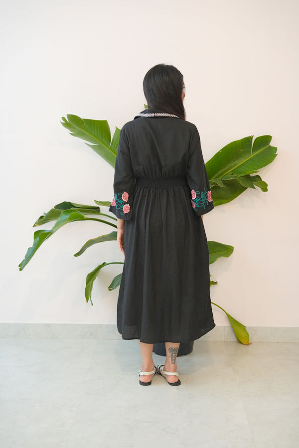 Person wearing a black dress with floral details standing between two potted plants.