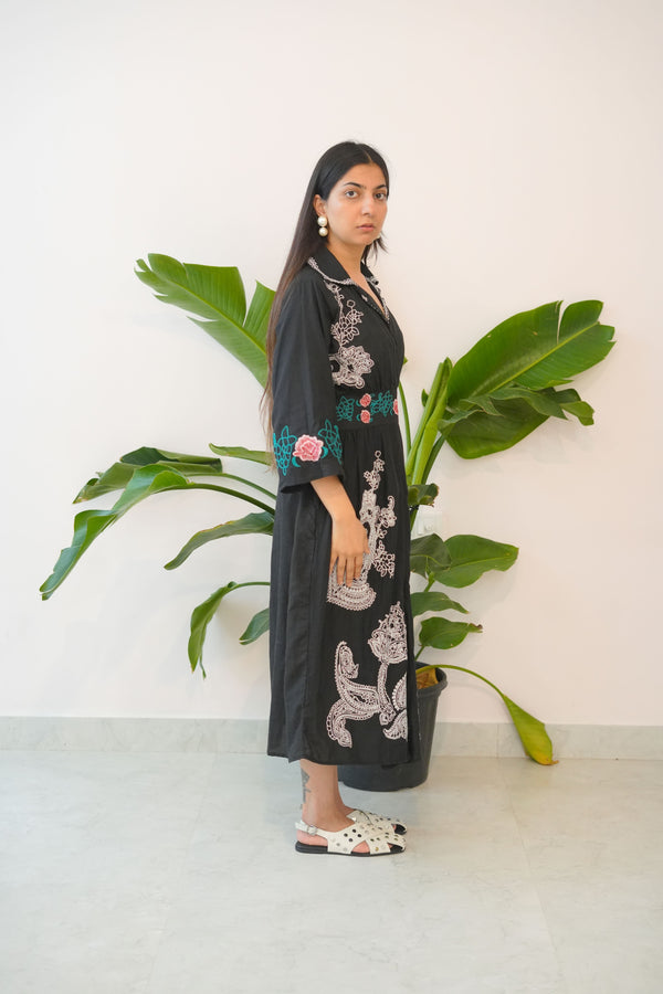 Woman in a long black dress with floral patterns standing next to a large green plant indoors.