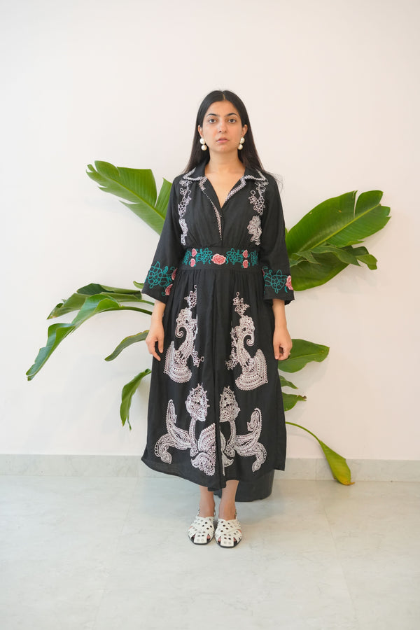Woman wearing a black dress with white embroidery standing in front of a plant.