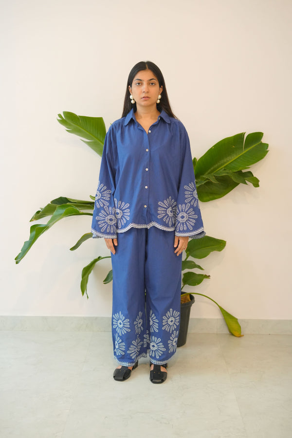 Woman wearing a blue outfit with white patterns standing against a white wall with green plants.