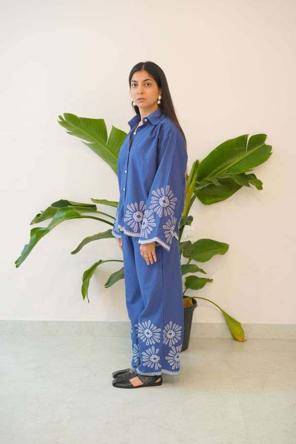Woman in a blue dress with floral patterns standing next to a large green leafy plant indoors.