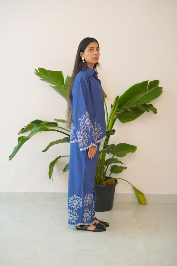 Woman in a blue outfit with white patterns standing next to a large green plant indoors.