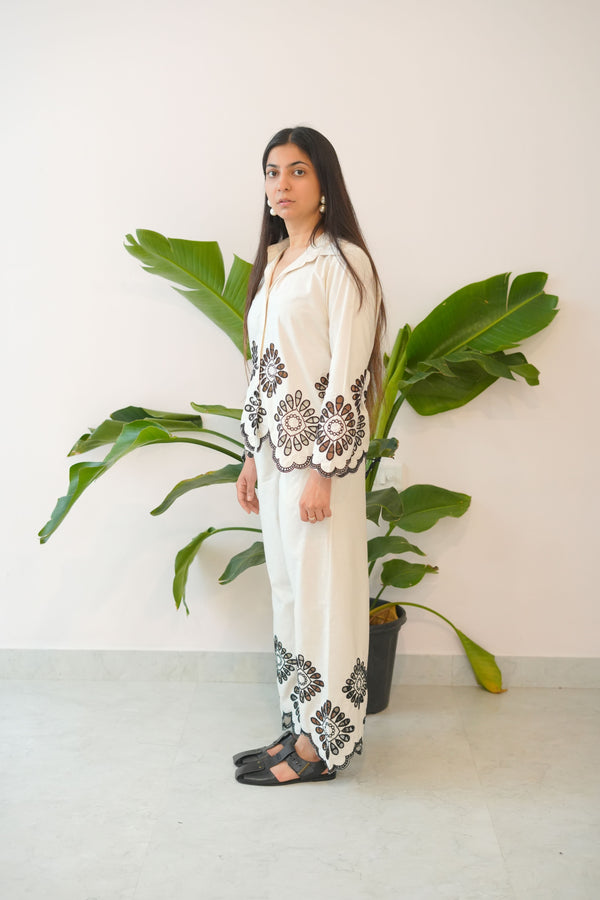 Woman in a white outfit with black lace details standing next to a large green plant against a white wall.
