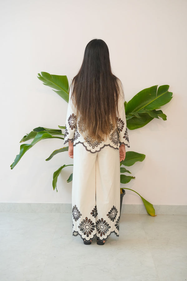 Person wearing a white dress with black lace details standing behind green leaves.