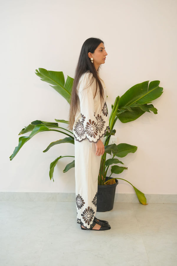 Person wearing a long-sleeve top with lace details and white pants, standing next to a large green leafy plant.
