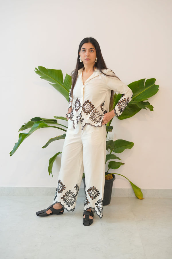 Woman in a white outfit with black patterns standing next to a plant against a white wall.