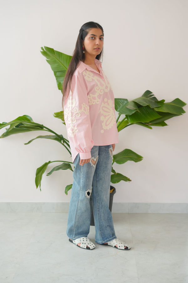Person wearing a pink top with floral patterns and blue jeans standing next to a plant indoors.