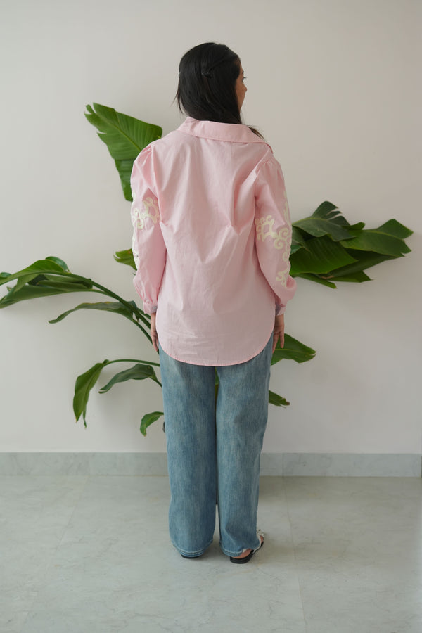 Person wearing a pink top and blue jeans standing next to a plant indoors.