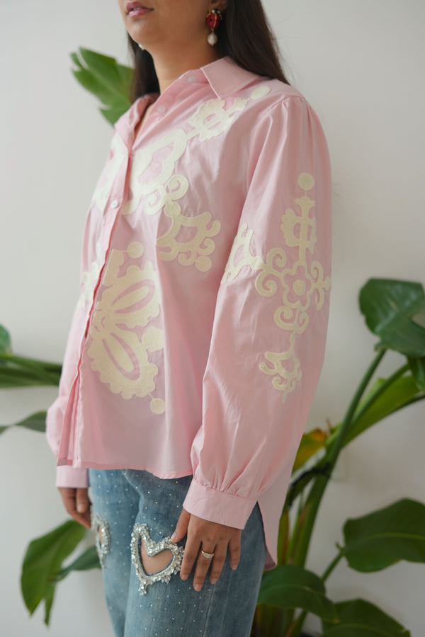 Person wearing a pink outfit with gold patterns, lying on a white surface with green plants around.