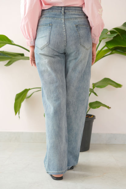 Person wearing blue jeans and a pink shirt standing against a white wall with green plants.
