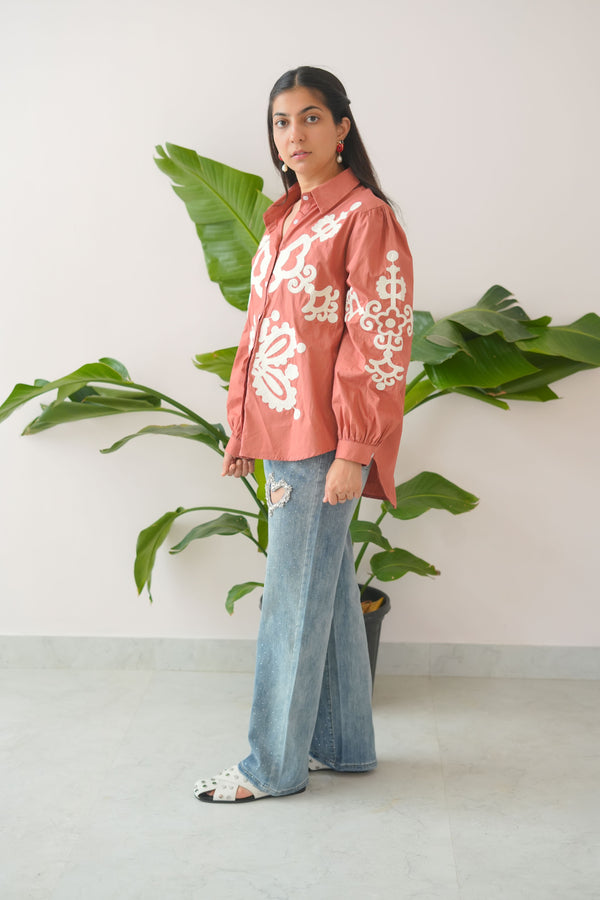 Person wearing a pink embroidered jacket and blue jeans standing in front of green plants.