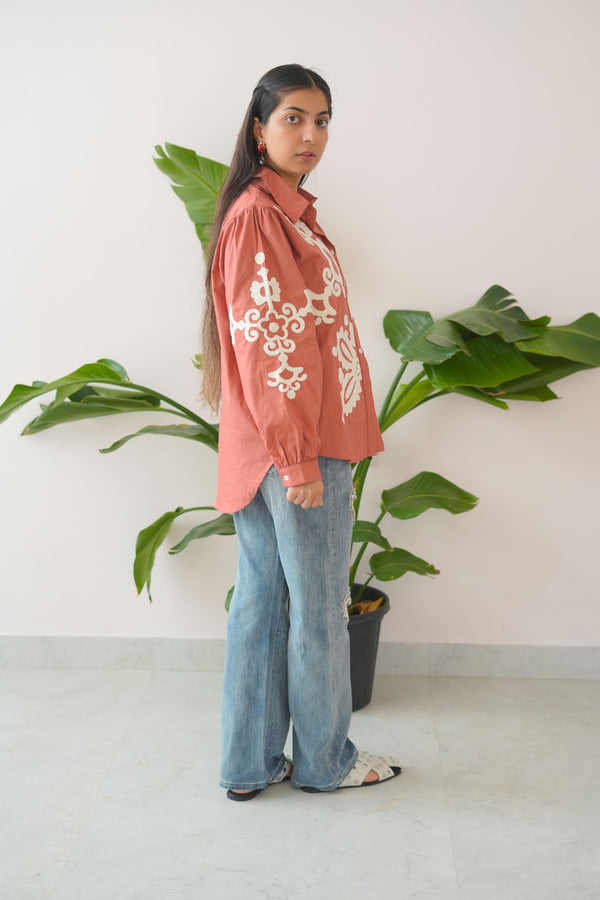 Person wearing a red hoodie with white patterns and blue jeans standing against a white wall with green plants.