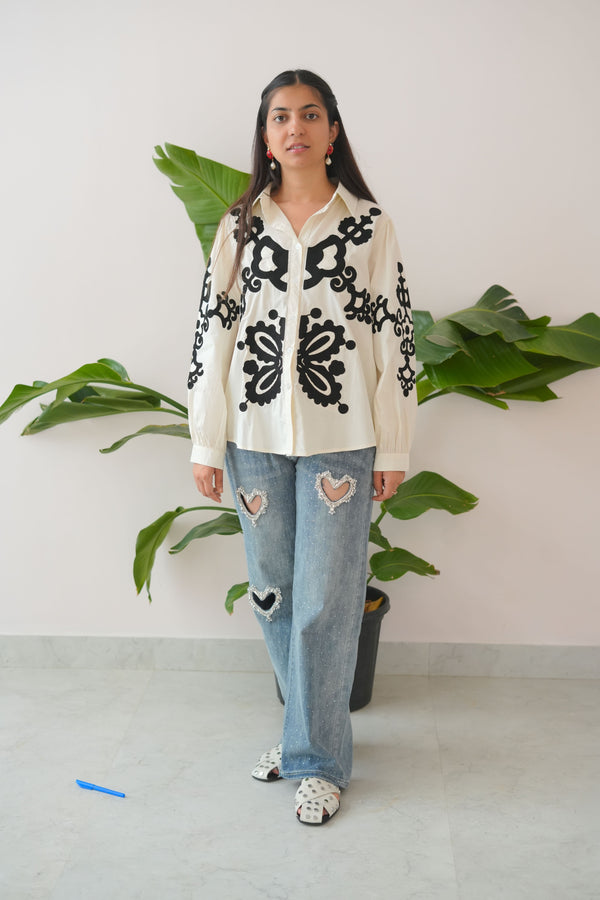 Person wearing a patterned top and jeans standing in front of green plants.