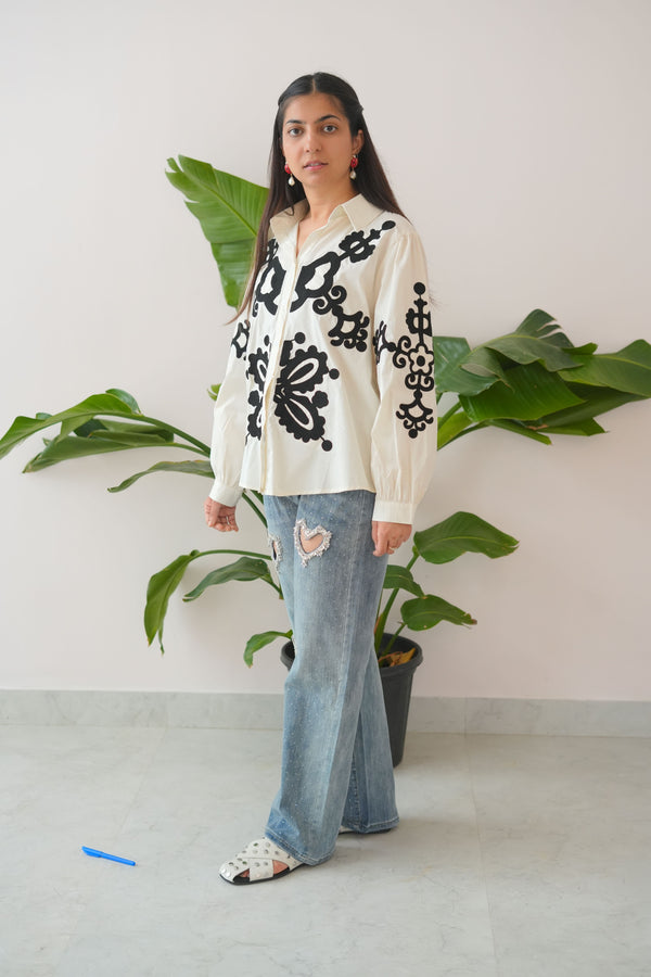 Person wearing a white cardigan with black embroidery and blue jeans standing in front of green plants.