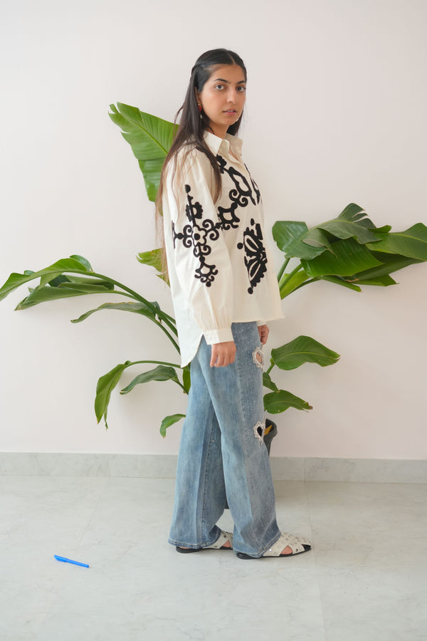 Person wearing a white sweater with black patterns and blue jeans standing in front of green plants.