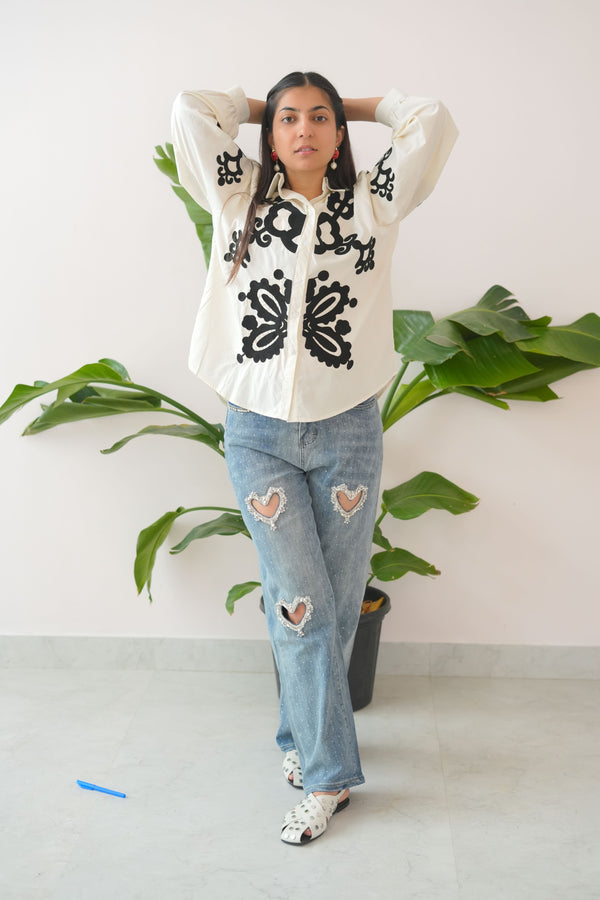 Person wearing a white dress with black patterns and blue jeans, lying on a white floor with green leaves.