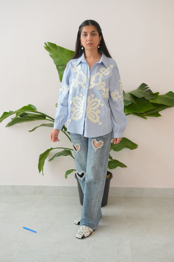 Person wearing a light blue traditional outfit with white embroidery, standing against a neutral background.