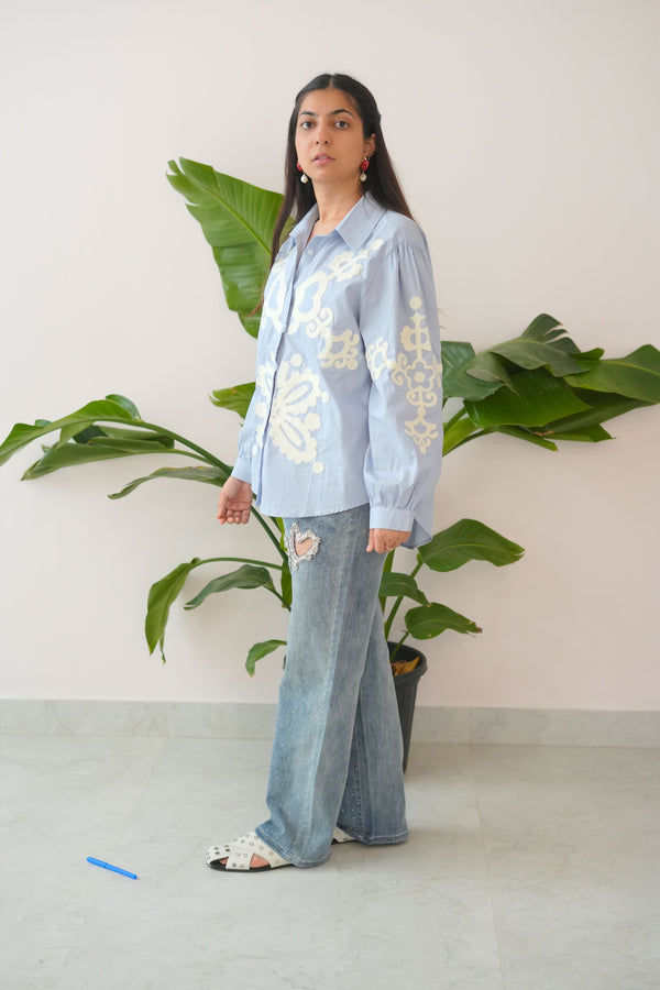 Woman in a light blue embroidered top and jeans standing in front of green plants.