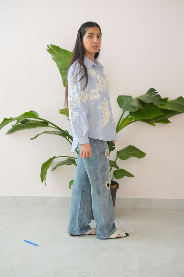 Person wearing a light blue outfit with floral patterns standing in front of a plant.
