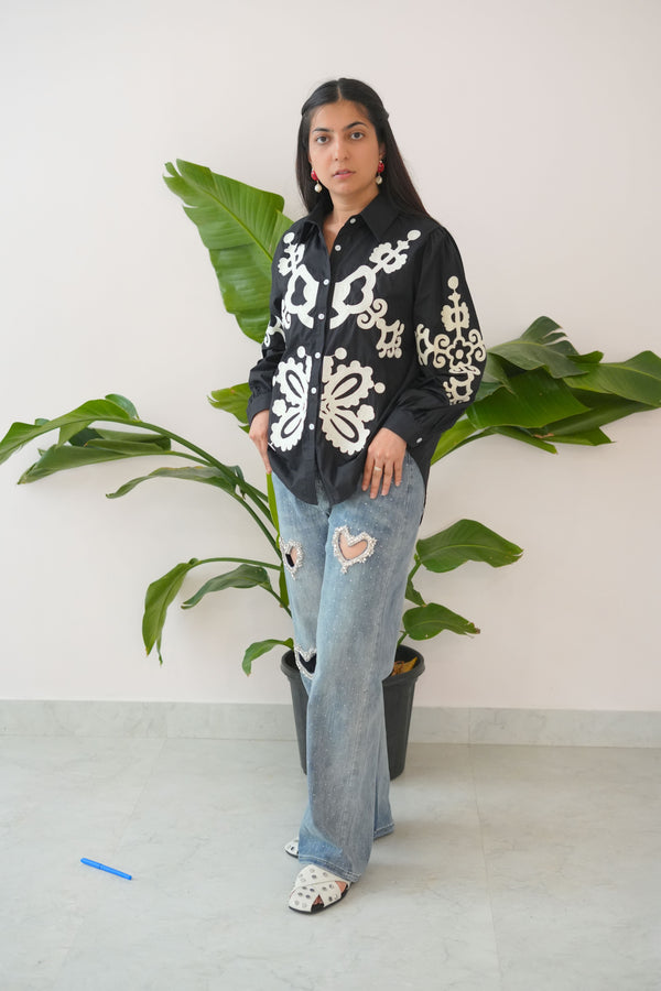 Person wearing a black patterned sweater and blue jeans standing against a white wall with green plants.