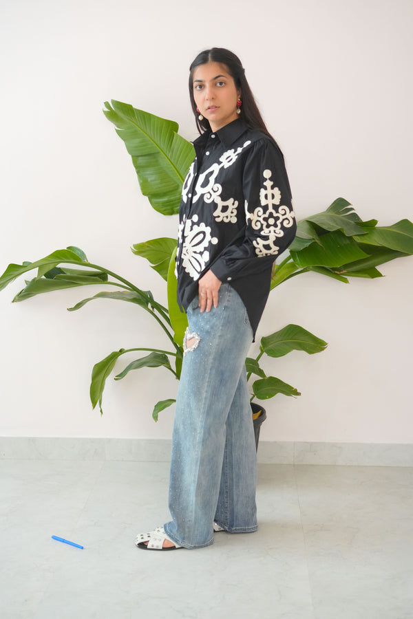 Person wearing a black and white patterned top with blue jeans standing next to a plant.