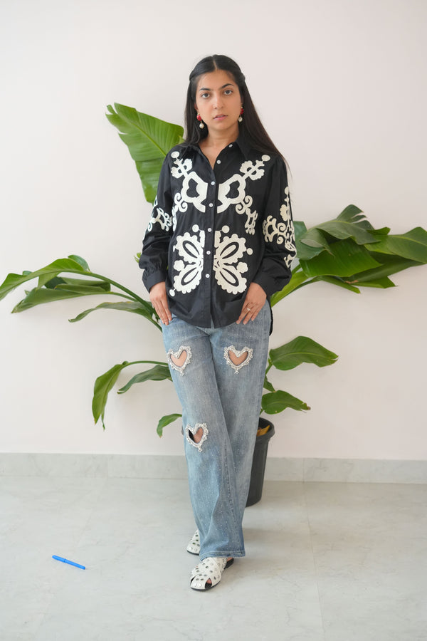 Person wearing a black hoodie with white patterns and ripped jeans standing against a white wall with green plants.