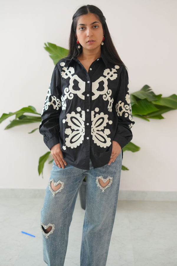 Person wearing a black hoodie with white patterns and ripped jeans standing against a white wall with green plants.