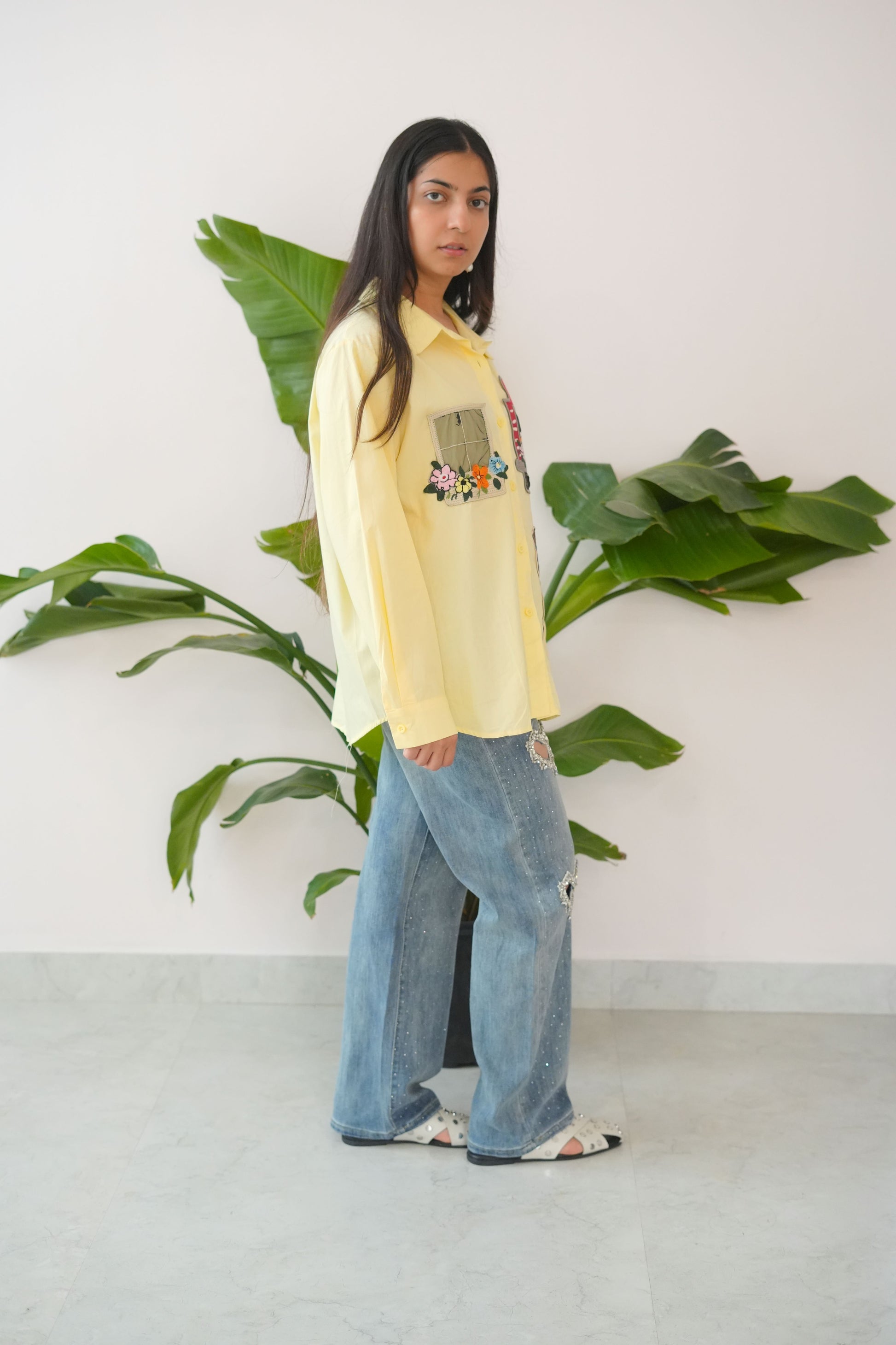 Person wearing a yellow sweatshirt and blue jeans standing next to a plant indoors.