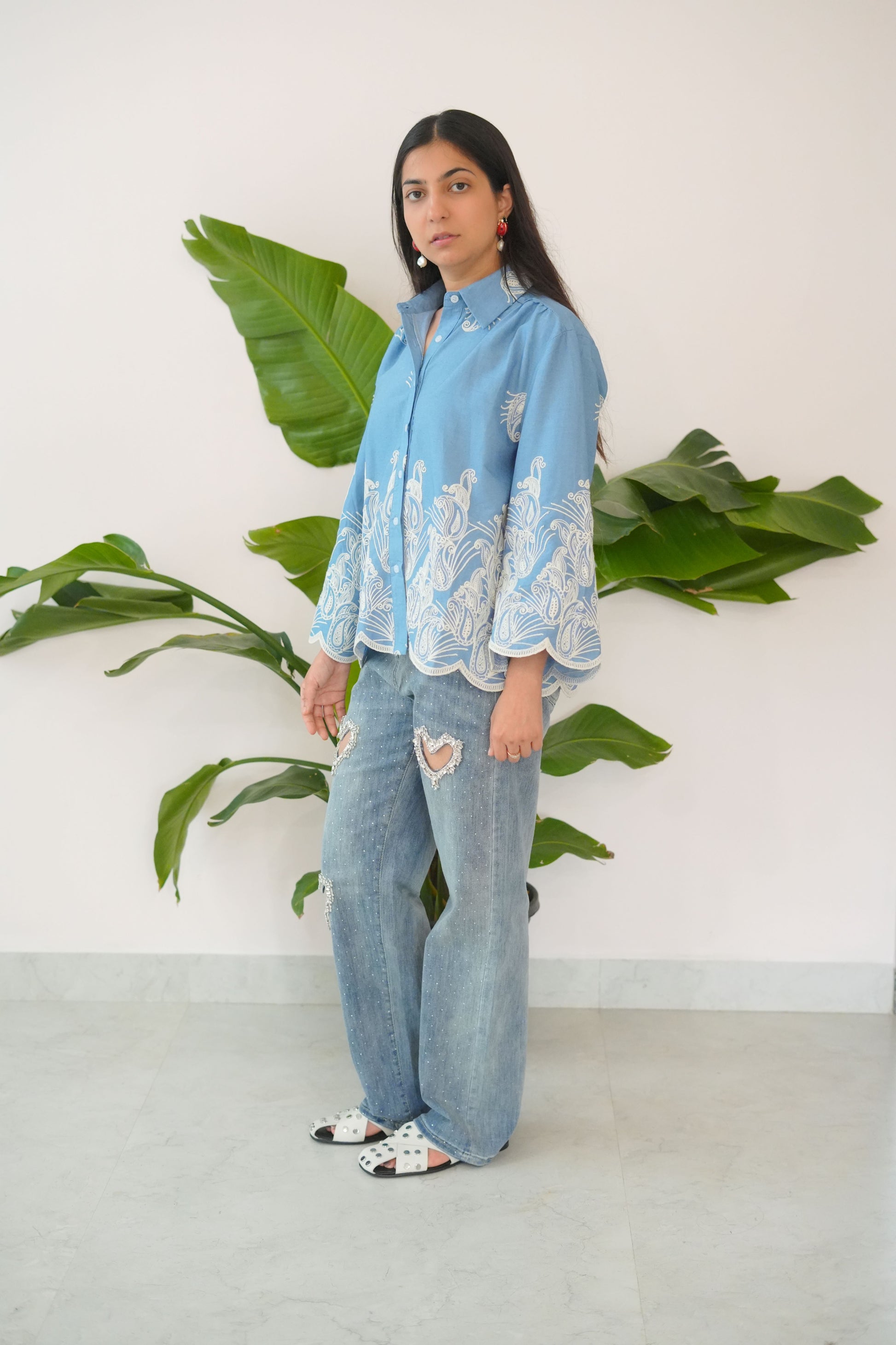 Person wearing a light blue embroidered top and ripped jeans standing among green plants.