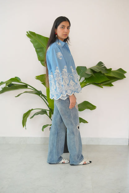Person wearing a blue denim outfit with white lace top, standing next to a large green plant indoors.