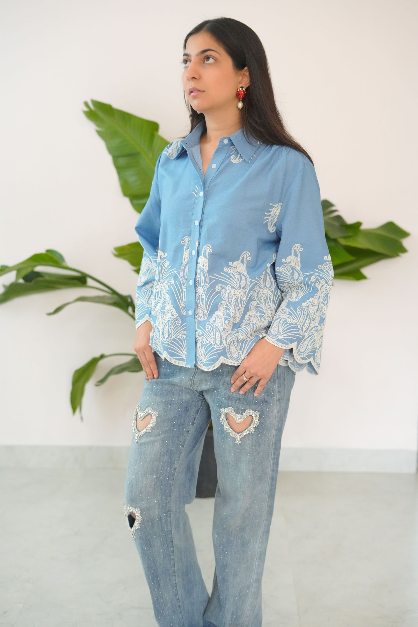 Woman wearing a light blue embroidered top and ripped jeans standing against a white background with green plants.