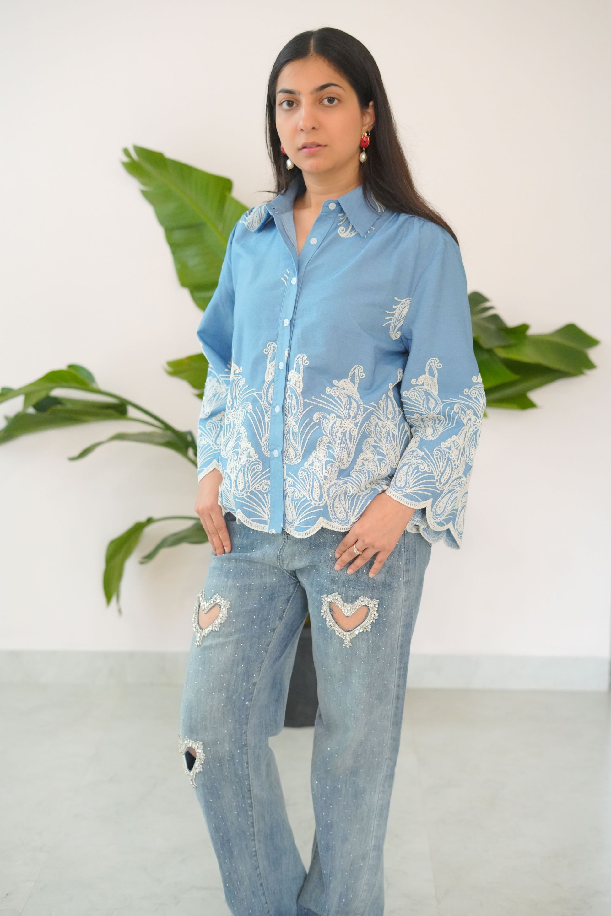 Woman wearing a blue embroidered top and ripped jeans standing against a white background with green plants.