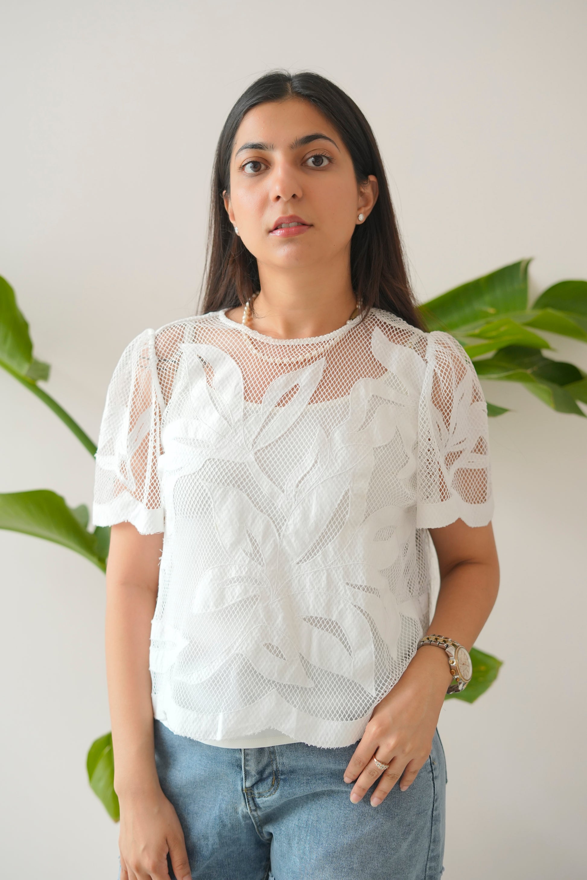 Woman wearing a white lace top and blue jeans standing against a light background with green plants.
