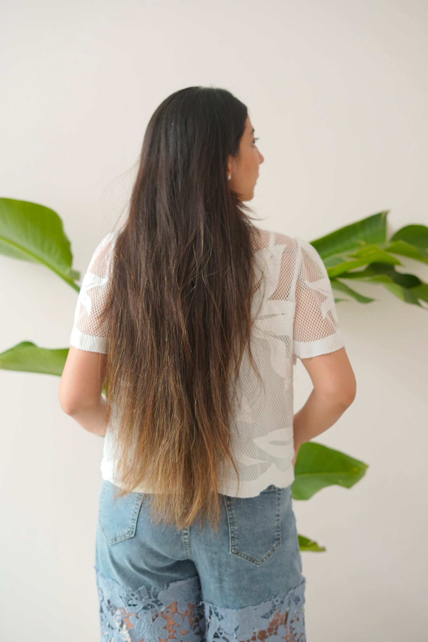 Person with long hair wearing a white lace top and blue jeans, standing against a white wall with green plants.