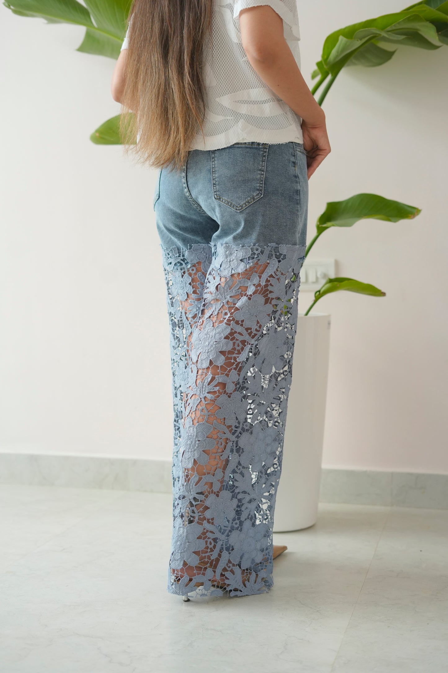 Person wearing light blue lace pants standing next to a white vase with green leaves.