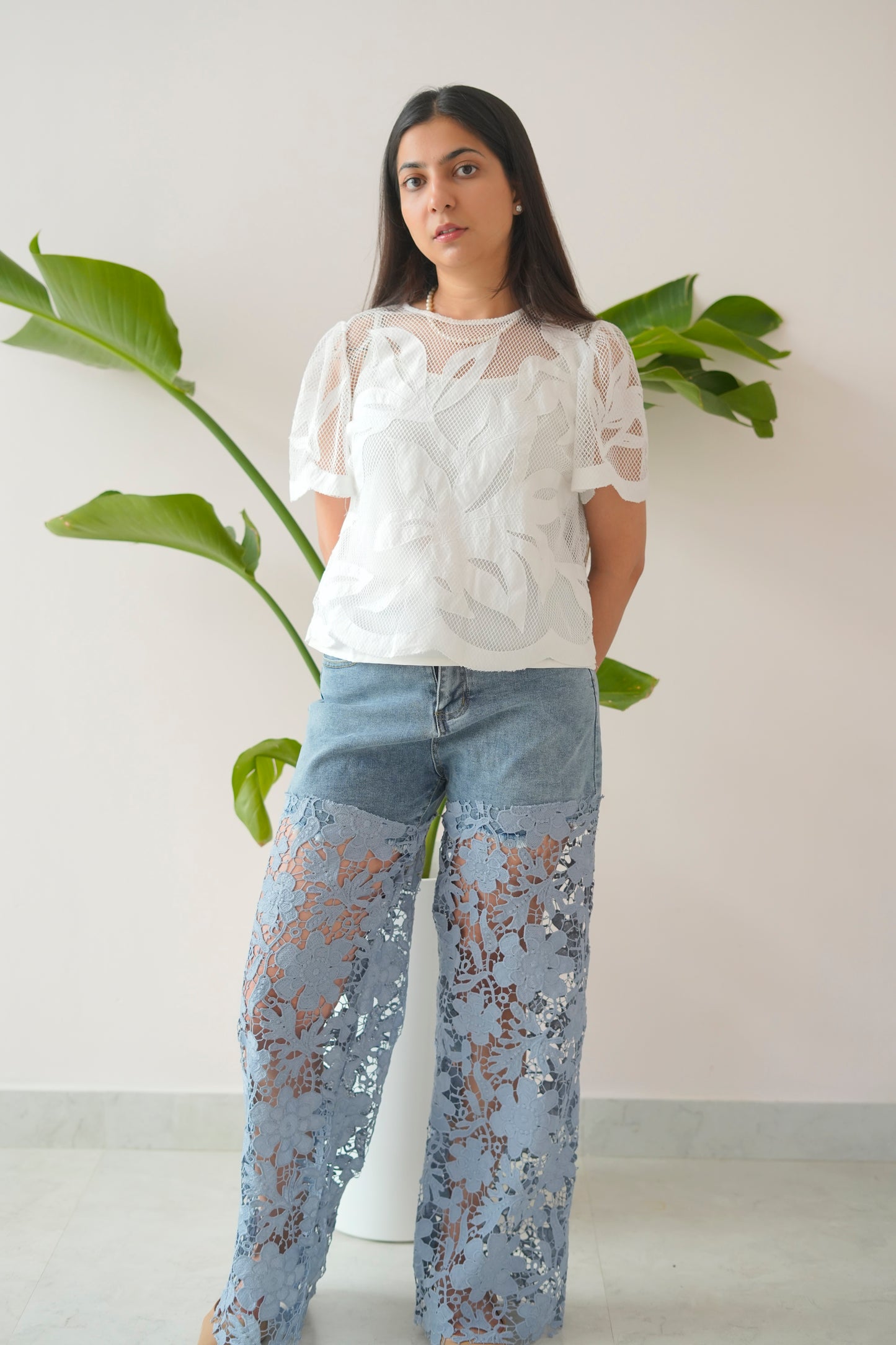 Woman wearing a white lace top and light blue jeans with floral patterns, standing against a plain background with a plant.