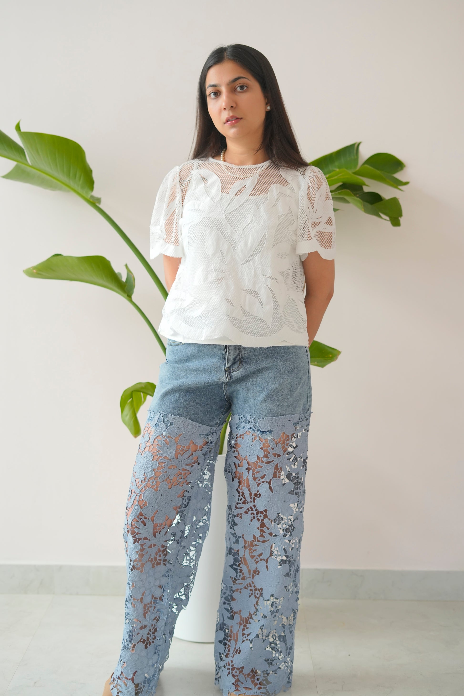 Woman wearing a white lace top and light blue jeans with floral patterns, standing against a plain background with a plant.