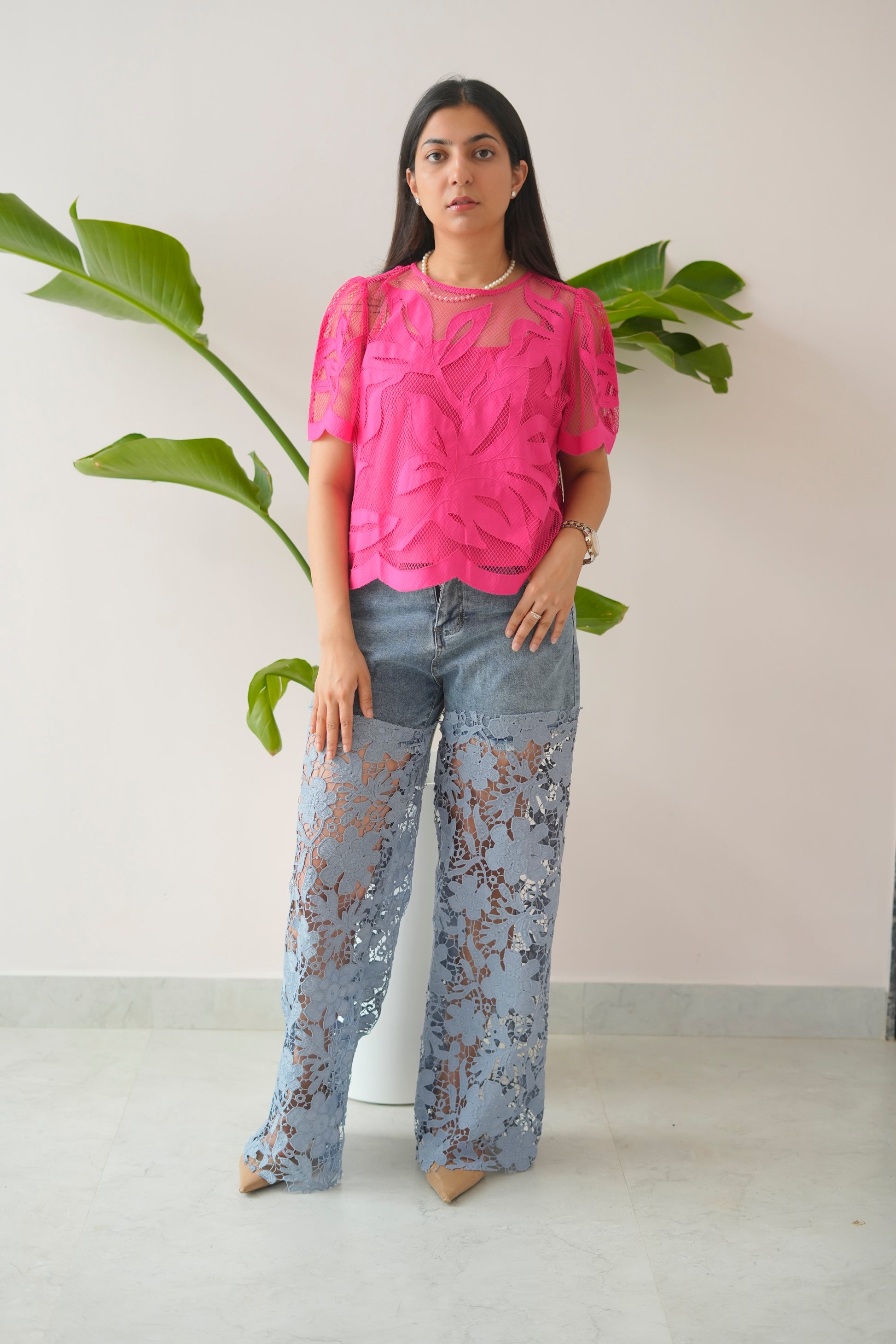 Woman wearing a pink top and light blue lace pants standing in front of a plant.