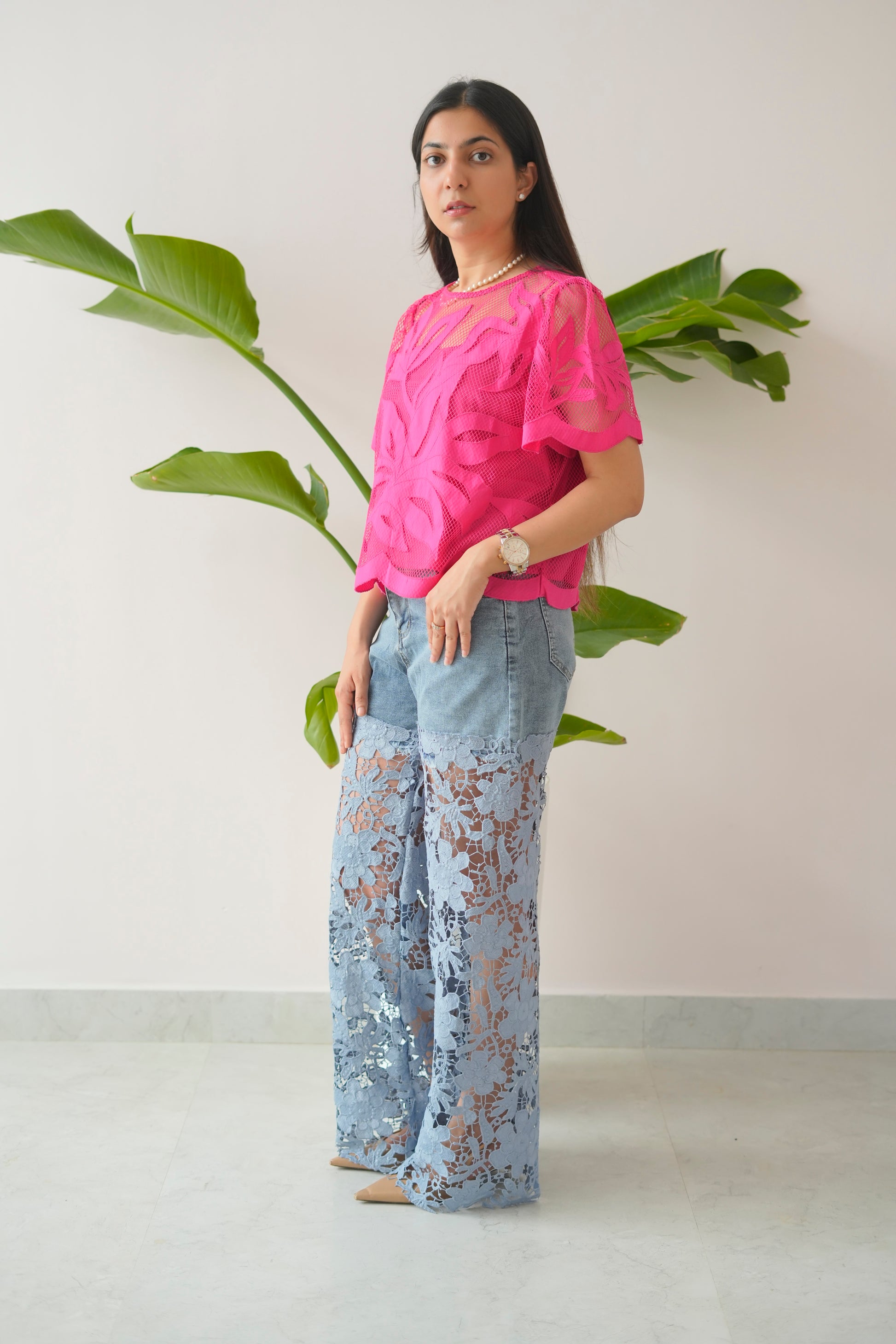 Woman wearing a pink top and light blue lace pants standing against a white wall with green plants.