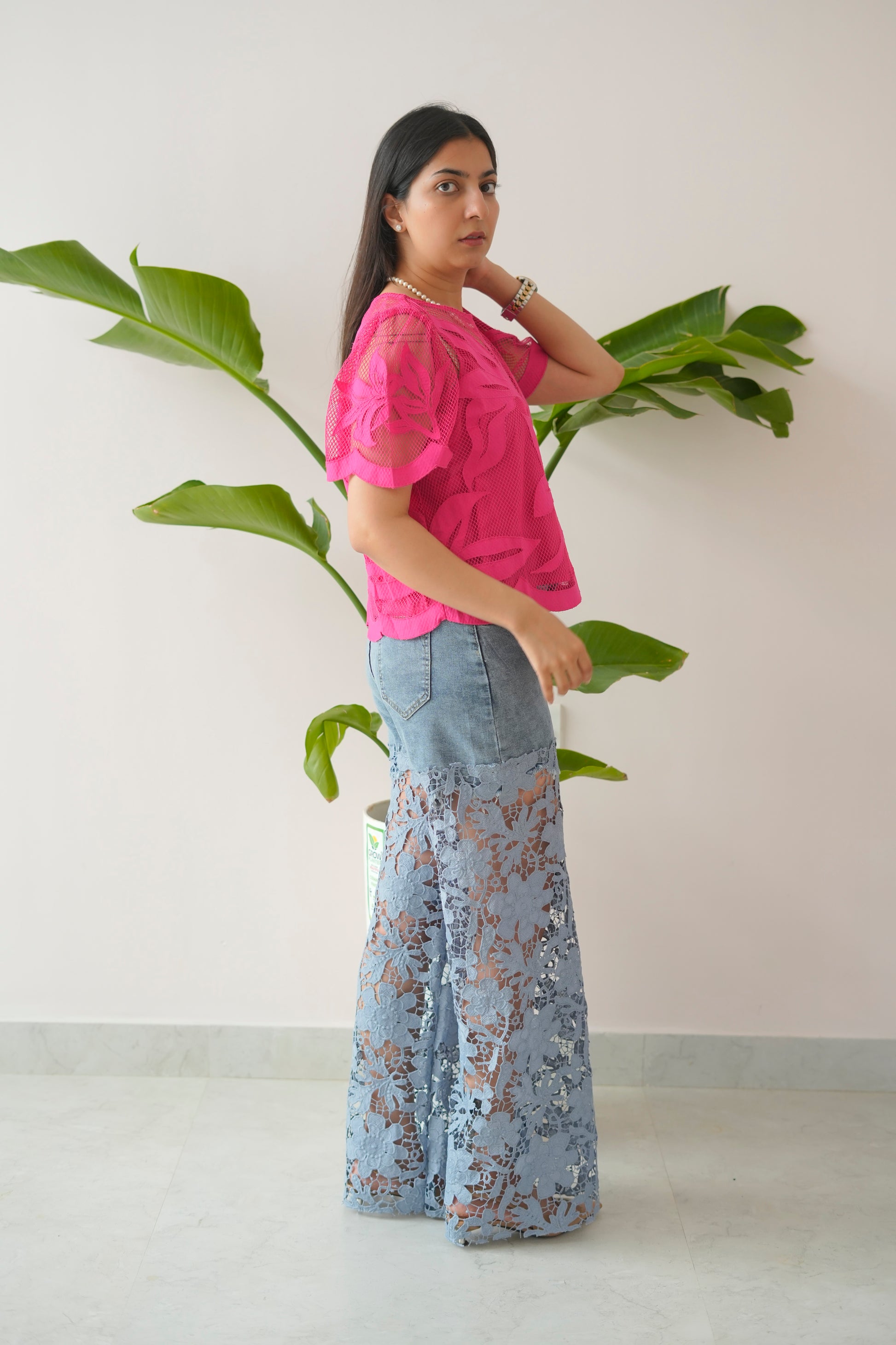 Woman wearing a pink top and light blue floral skirt standing next to a plant against a white wall.