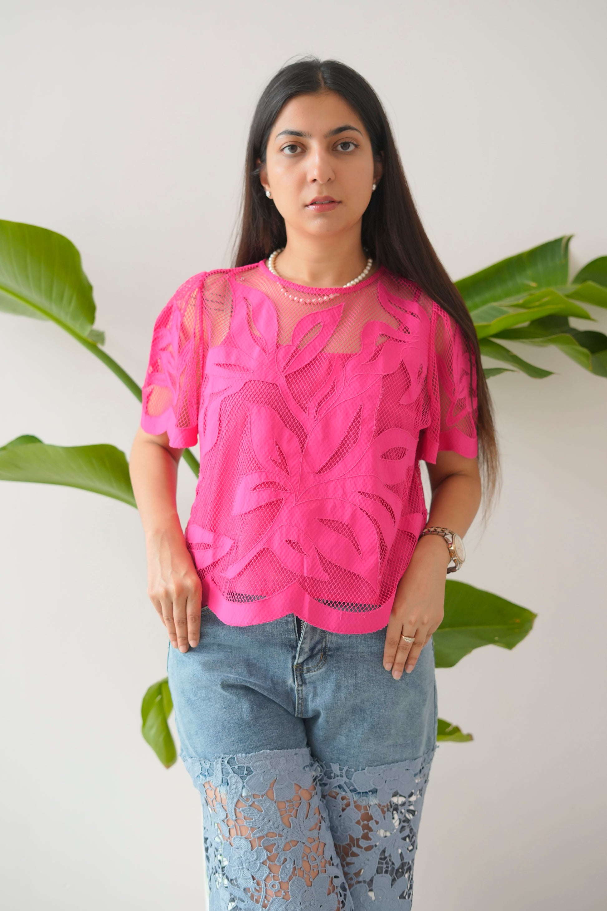 Woman wearing a bright pink top with floral patterns and light blue jeans against a white background with green leaves.
