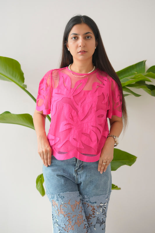 Woman wearing a bright pink top with floral patterns and light blue jeans against a white background with green leaves.